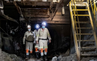 Three men with headlamps on are walking through a decrepit mine shaft