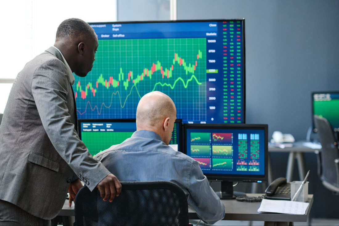 Two professional men are looking at multiple screens displaying trading graphs.