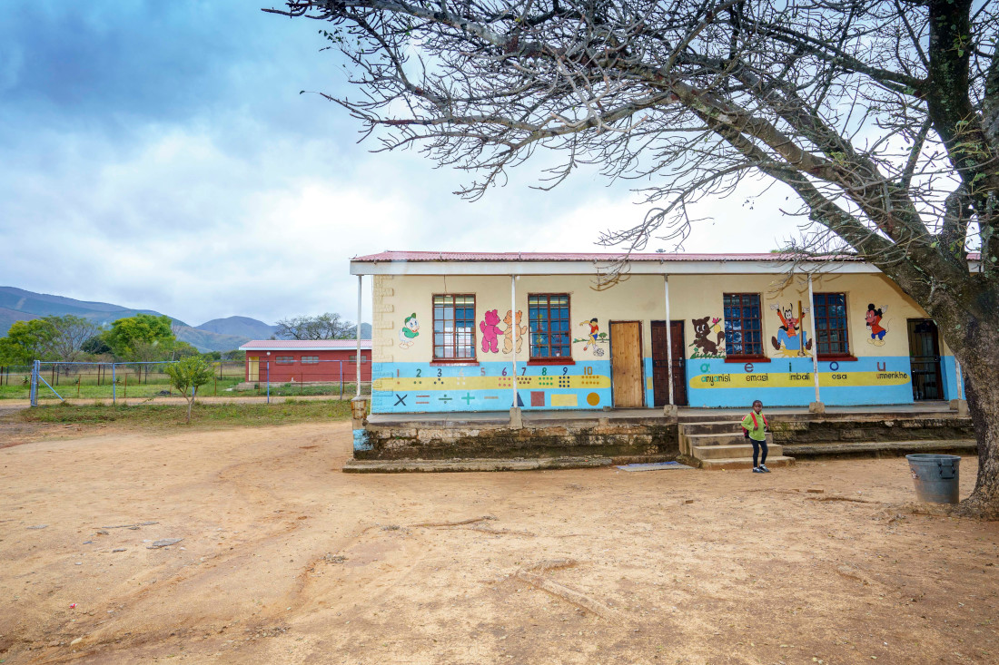 A colourful school building with greenery in the background and a young learner standing in front of it.